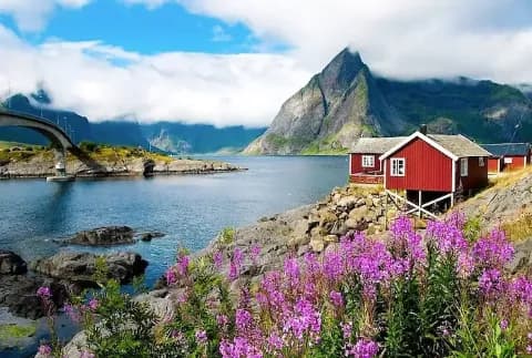 Ein rotes Fischerhaus aus Holz steht direkt am Wasser in einem bergigen Fjord in den Lofoten in Norwegen.
