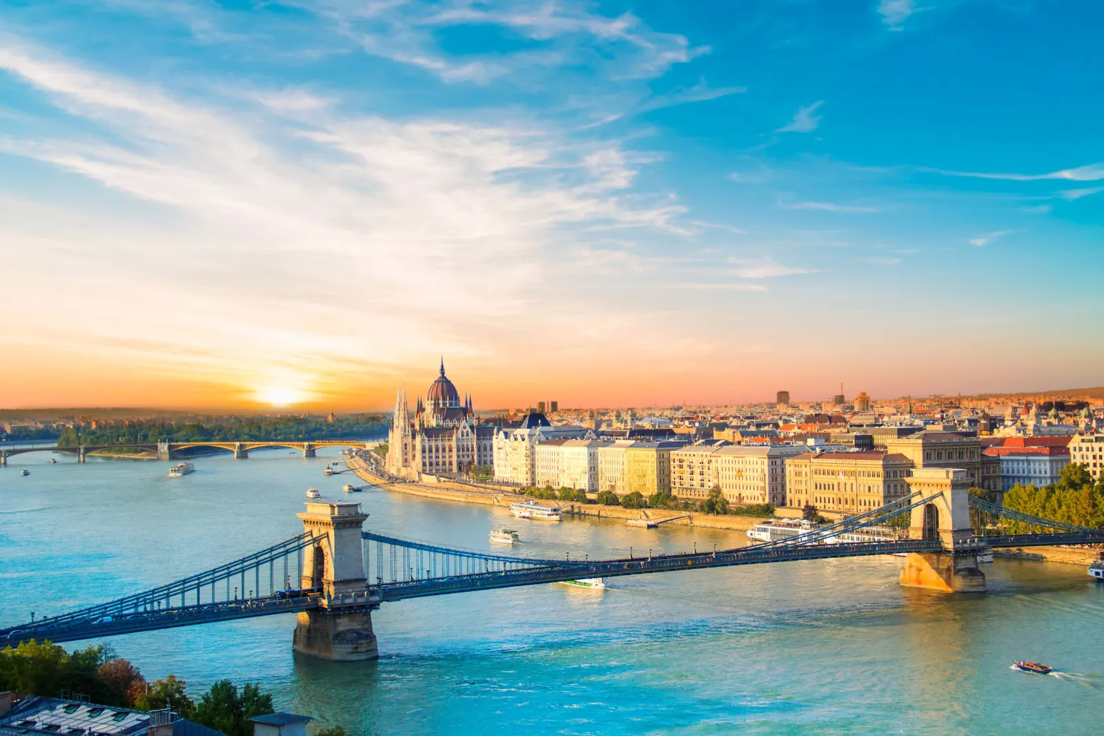 Der Blick richtet sich vom Fluss aus auf die Stadt Budapest in Ungarn, wobei eine Brücke über dem Fluss und alte Häuserfassaden am Stadtrand hin zum Fluss zu sehen sind.