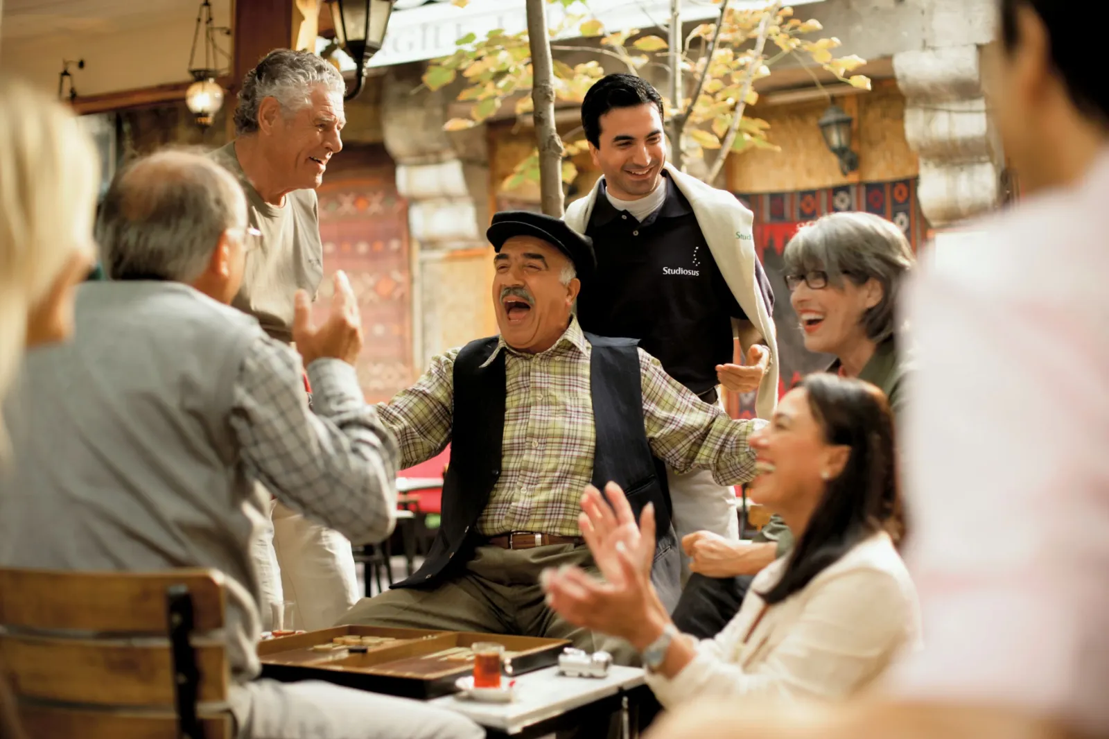 Auf dem Bild ist ein Studiosus-Reiseleiter mit seiner Gruppe in der Türkei bei einer Begegnung mit einem Einheimischen zu sehen, der gerade Backgammon gegen einen älteren Herrn spielt und herzlich lacht.