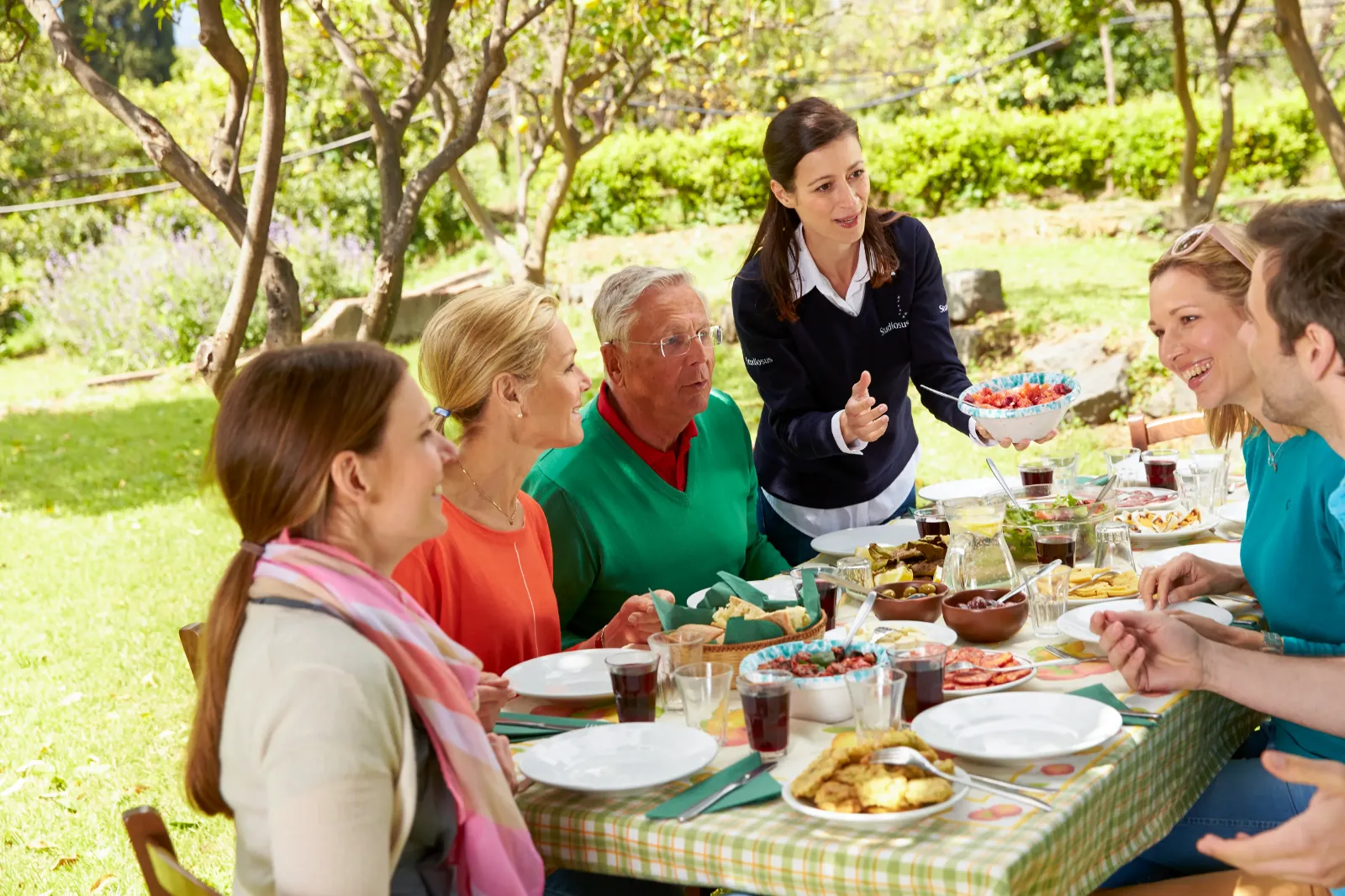 Auf dem Bild ist ein Tisch draußen im Grünen voll mit Essen zu sehen, an dem fünf Personen sitzen und eine Studiosus-Reiseleiterin steht, die Essen reicht und etwas dazu erklärt.