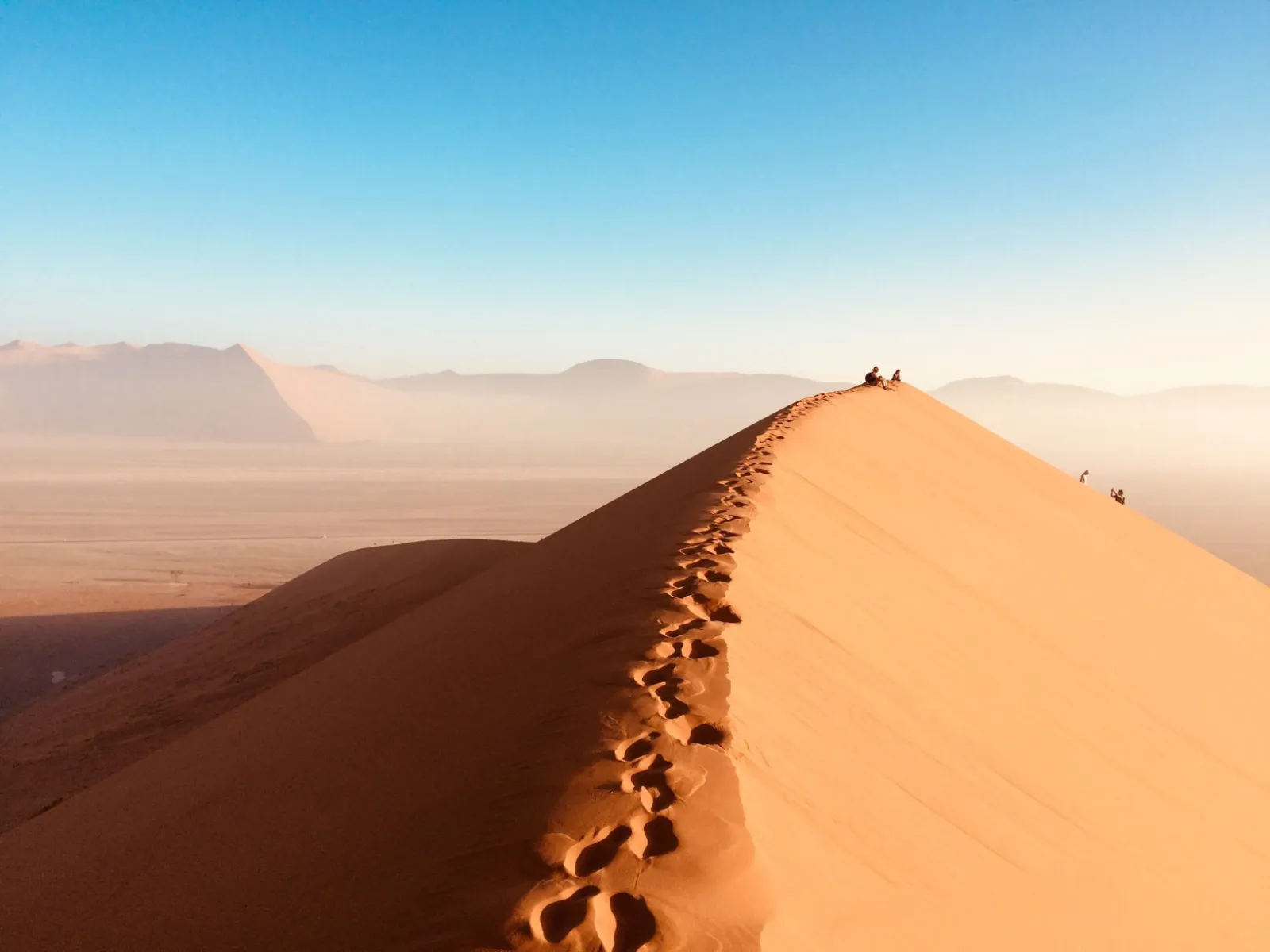 Auf dem Foto ist eine Düne in der Wüste abgebildet, auf dessen oberer Kante Fußspuren im Sand zu sehen sind, während im Hintergrund auch einige Personen unterwegs sind.