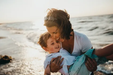 Eine Mutter, die glücklich ihr Kind am Strand hält, während die Wellen im Hintergrund rauschen.