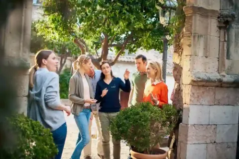 Eine Reisegruppe steht im Freien unter Bäumen und hört eine Reiseleiterin zu.