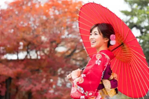 Eine junge japanische Frau in einem roten Kimono mit einem roten Sonnenschirm steht in einem Park.