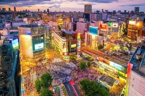 Ein Panoramablick von oben auf der berühmten Shibuya-Kreuzung im abendlichen Tokio mit vielen Menschen auf mehreren Zebrastreifen zwischen Hochhäusern mit Leuchtreklamen.