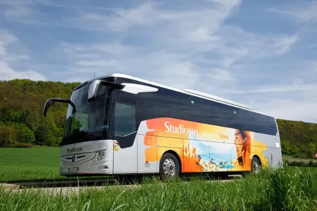 Ein Studiosus Reisebus auf einer Straße, umgeben von üppigem Grün und sanften Hügeln.