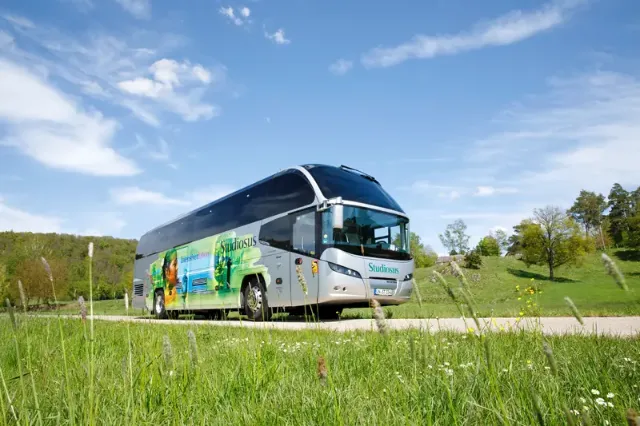 Ein moderner Reisebus von Studiosus fährt durch eine grüne Landschaft unter blauem Himmel.