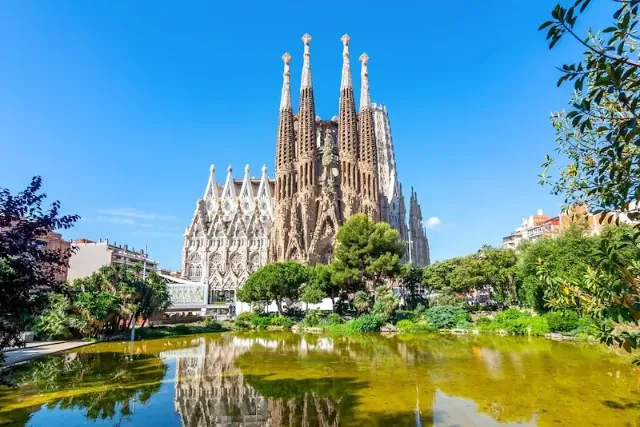 Die Sagrada Familia Kathedrale in Barcelona an einem Park mit Teich und bei blauem Himmel.