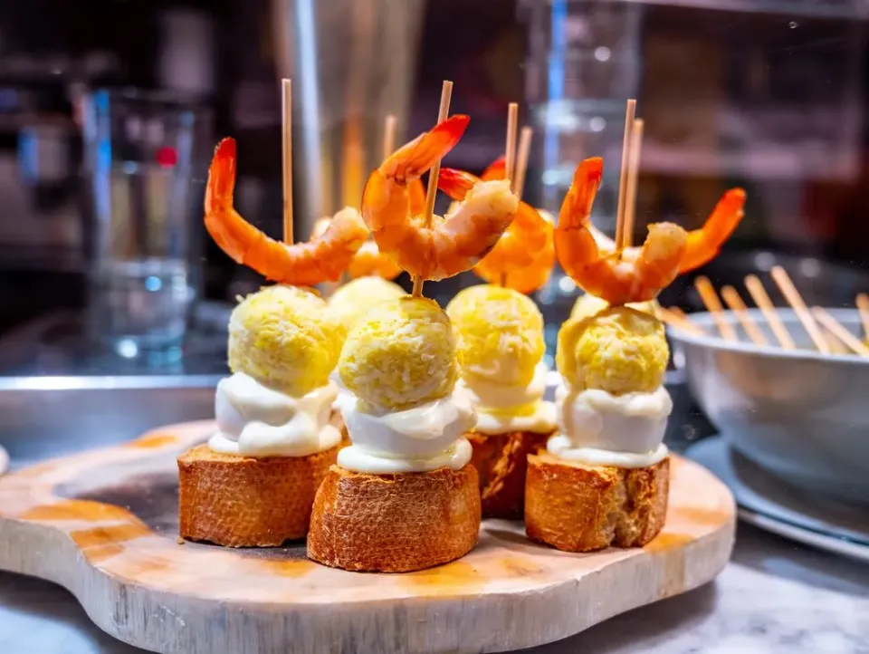 Ein Holzbrett mit appetitlichen Pintxos, ein typischer Snack in den Bars des Baskenlandes, kleine Brotscheiben, die mit verschiedenen Zutaten belegt sind und geschmackvoll hergerichtet werden.

