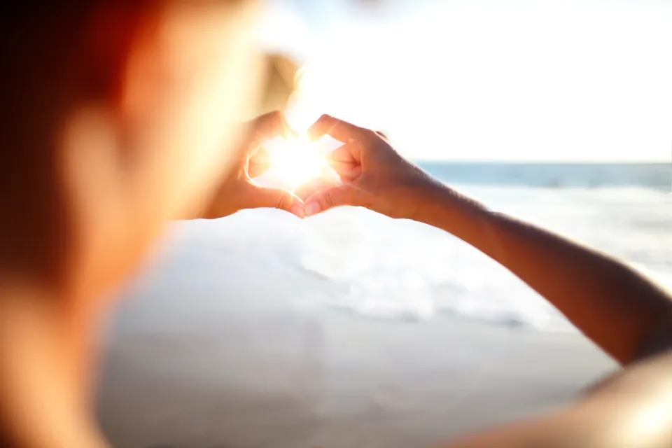 Hände formen ein Herz, das die untergehende Sonne umschließt.