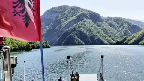 Bootstour auf einem See in Albanien mit Blick auf die bewaldeten Hügel. Die albanische Flagge ist im Vordergrund sichtbar.