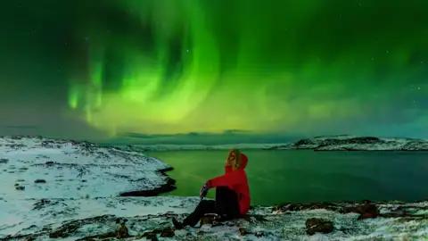 Eine Person in roter Winterjacke sitzt auf einem schneebedeckten Felsen und beobachtet die grünen Nordlichter am Himmel. Mit etwas Wetterglück bestaunen auch Sie auf dieser Reise Polarlichter.