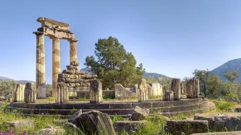 Auf unserer Gruppenreise mit Studiosus bewundern wir die Tholos von Delphi, ein zentraler Besichtigungspunkt, welchert ihre charakteristischen Säulen in einer grünen Landschaft unter blauem Himmel zeigt.