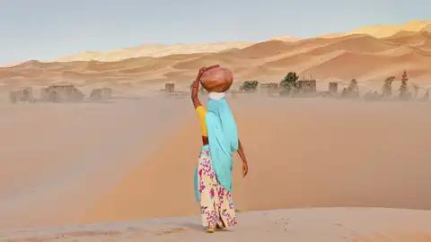 Eine Frau in traditioneller Kleidung transportiert einen Wasserkrug durch die Wüste, was einen Einblick in das Leben der Einheimischen gibt, den wir auf unserer Gruppenreise mit Studiosus bekommen.