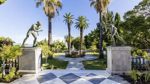 Bronzestatuen im sonnigen Schlossgarten von Sisis Achilleion-Palast auf Korfu, umgeben von hohen Palmen und grünen Pflanzen, ein Highlight unserer Studiosus Gruppenreise.