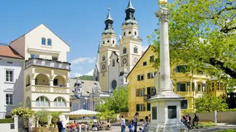 Der barocke Brixner Dom an einem sonnigen Tag, gesehen vom belebten Domplatz mit seinen Cafés und historischen Gebäuden, ein toller Ausblick auf unserer Studiosus Gruppenreise.