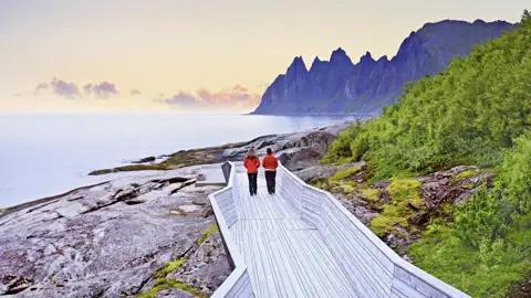 Ein Paar in roten Jacken auf einem Holzsteg an der Felsküste von Senja, im Hintergrund die Okshornan-Gipfel, ein tolles Naturhighlight auf userer Gruppenreise mit Studiosus.
