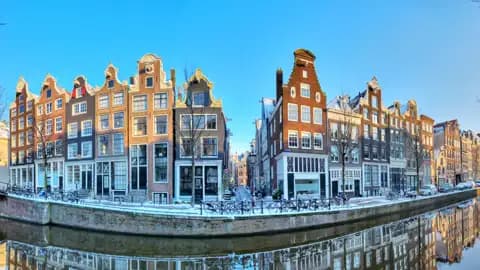 Typische Amsterdamer Giebelhäuser mit schneebedeckten Dächern spiegeln sich im ruhigen Wasser einer Gracht im Winter, ein Highlight auf unserer Eventreise in die Niederlande.