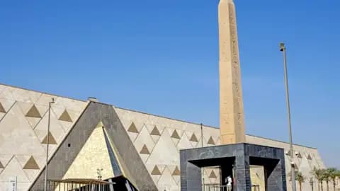 Die moderne Fassade des Grand Egyptian Museum in Gizeh, davor ein hoher, antiker Obelisk unter strahlend blauem Himmel, ein architektonisches Highlight unserer Gruppenreise.