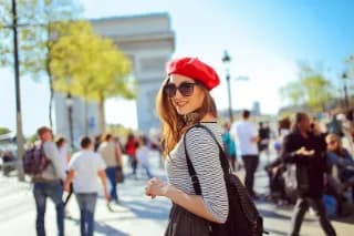 Eine lächelnde Frau mit Sonnenbrille, einer roten Mütze und modischer Kleidung auf den Straßen von Paris mit dem Arc de Triomphe im Hintergrund.