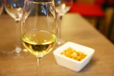 Nahaufnahme eines Weinglases mit hellem Wein und einer Schüssel Snacks im Hintergrund.