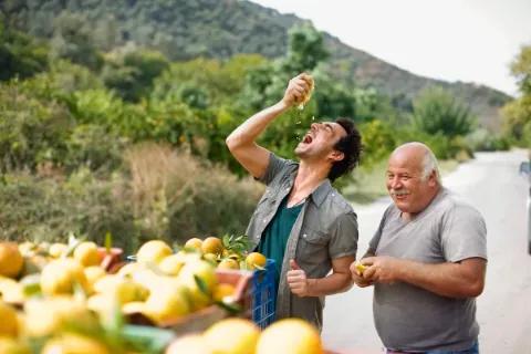 Ein Mann presst eine Orange und lacht dabei, während ein anderer zuschaut.