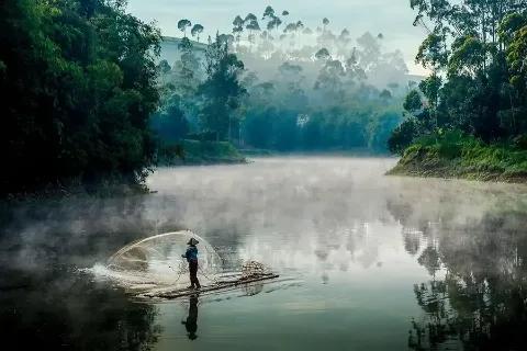Ein Fischer steht im Nebel am Ufer eines ruhigen Flusses.