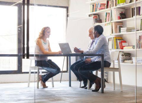 Drei Menschen sitzen in einem Raum mit Büchern vor einem Laptop.
