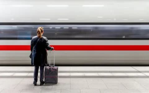 Eine Frau steht mit ihrem Koffer am Bahnsteig während ein ICE einfährt.