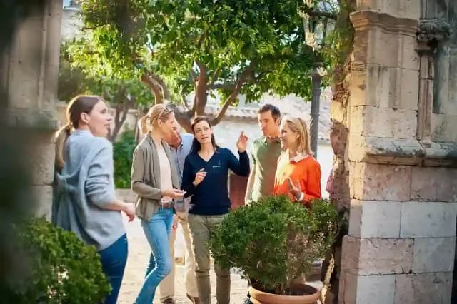 Eine Reisegruppe steht im Freien unter Bäumen und hört eine Reiseleiterin zu.