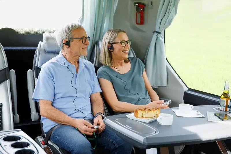 Ein älteres Paar genießt eine Reise in einem Reisebus mit Kopfhörern und Snacks.