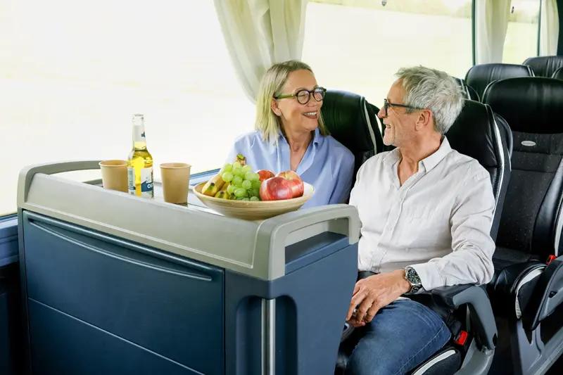 Ein älteres Paar genießt eine entspannte Busfahrt mit Obst und Getränken.