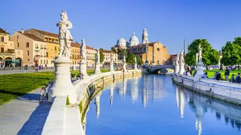 Auf unserer Rundreise nach Italien bewundern wir dernvon Statuen gesäumten Kanal am Prato della Valle in Padua mit der Basilika Santa Giustina im Hintergrund bei Sonnenschein.