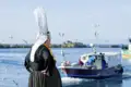 Frau in bretonischer Tracht am Hafen blickt auf ein Fischerboot.