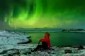 Eine Person in roter Winterjacke sitzt auf einem schneebedeckten Felsen und beobachtet die grünen Nordlichter am Himmel. Mit etwas Wetterglück bestaunen auch Sie auf dieser Reise Polarlichter.