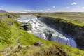 Eine Reisegruppe auf einem Wanderpfad am imposanten Gullfoss Wasserfall, der in eine grüne Schlucht in Island stürzt, ein Naturhighlight jeder Gruppenreise nach Island