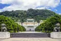Auf unserer Gruppenreise mit Studiosus werfen wir einen Blick auf den Eingangsbereich des Nationalen Palastmuseums in Taipeh, mit steinernen Wächterlöwen, einer breiten Treppe und dem Hauptgebäude vor einem grünen Hügel.