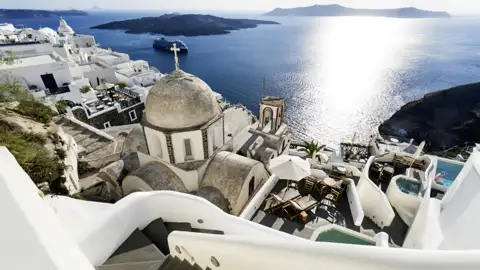 Weiße Gebäude und eine Kuppelkirche auf Santorin mit Blick auf das Meer und ein Kreuzfahrtschiff