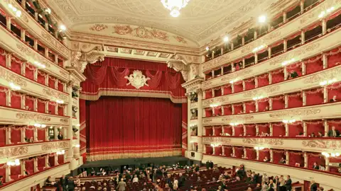 Auf unserer Gruppenreise mit Studiosus genießen wir die  Innenansicht der Mailänder Scala. Zu sehen ist der opulente Zuschauerraum mit Publikum in den Logen und im Parkett vor der Bühne mit dem roten Vorhang. Ein Highlight jeder Eventreise nach Mailand.