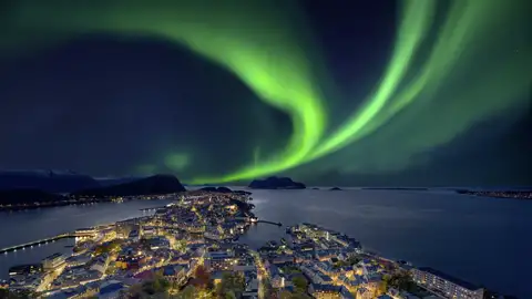 Grüne, tanzende Nordlichter am Sternenhimmel über der beleuchteten Küstenstadt Ålesund und der Fjordlandschaft Norwegens auf ihrer Studiosus Eventreise.