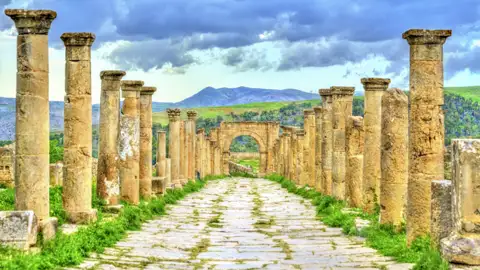 Gepflasterte Säulenstraße in der antiken Römerstadt Djemila, UNESCO-Welterbe in Algerien, mit grünen Bergen im Hintergrund, Auf ihrer Studiosus Eventreise zu besichtigen.