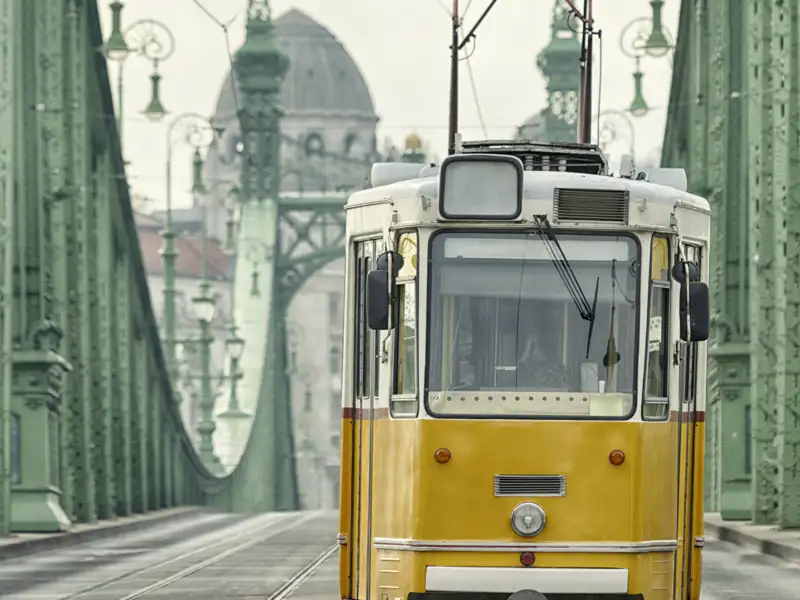 Eine ikonische gelbe Straßenbahn überquert die grüne Freiheitsbrücke in Budapest an einem bewölkten Tag auf einer Eventreise.