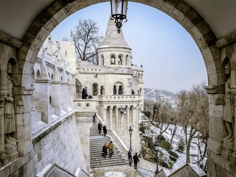 Blick durch einen steinernen Torbogen auf die winterliche Fischerbastei im Burgviertel von Budapest mit Besuchern auf den Treppen. Sie besuchen die Fischerbastei während der Studienreise.