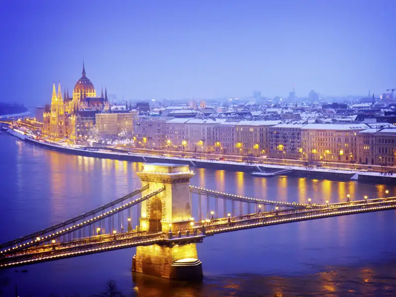 Blick auf die beleuchtete Kettenbrücke über der Donau und das verschneite Parlamentsgebäude in Budapest bei Dämmerung.
