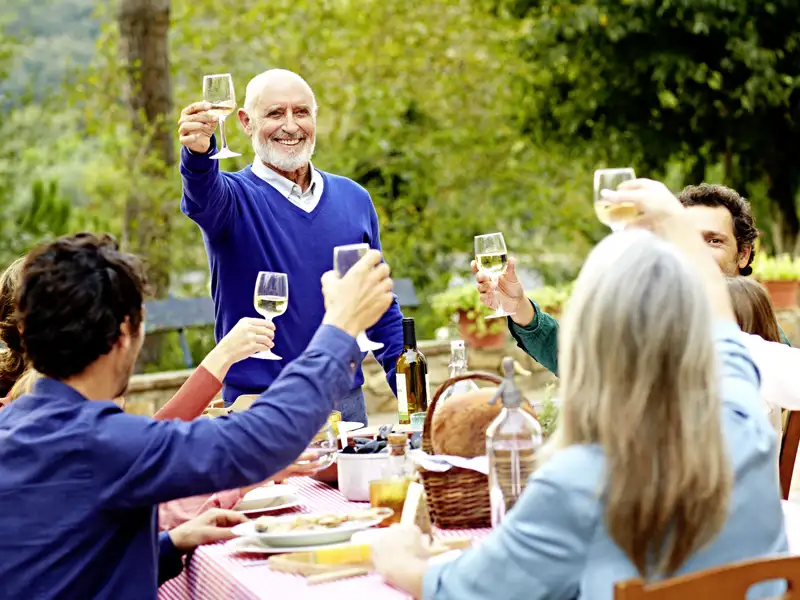 Eine Gruppe von Freunden sitzt an einem Tisch im Freien und genießt eine Mahlzeit. Sie heben ihre Weingläser zu einem Toast.