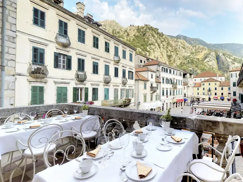 Aussicht von dem Balkonrestaurant unseres Hotels in Kotor auf einen belebten Marktplatz, umgeben von historischen Gebäuden und Bergen