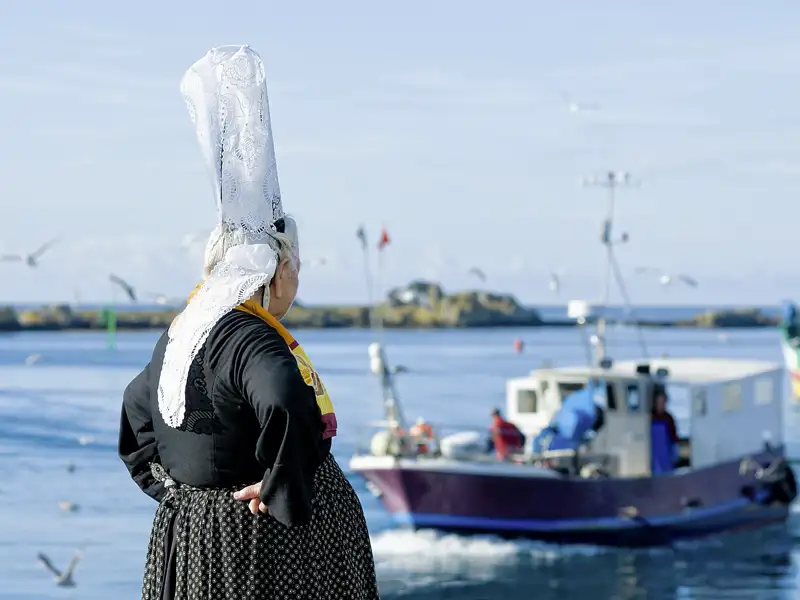 Frau in bretonischer Tracht am Hafen blickt auf ein Fischerboot.