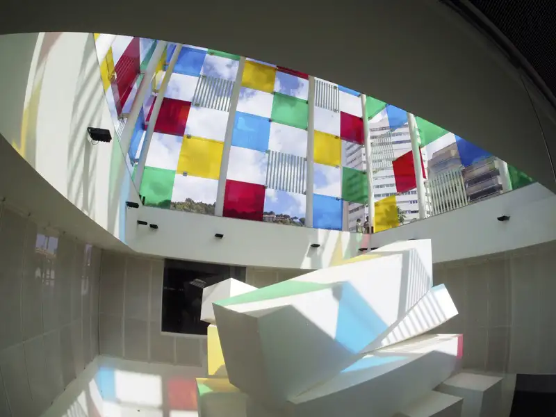 Innenansicht des Centre Pompidou in Málaga mit seiner bunten Glasdecke, die als Kubus geformt ist. Das bunt gefärbte Licht fällt auf eine weiße moderne Skulptur.