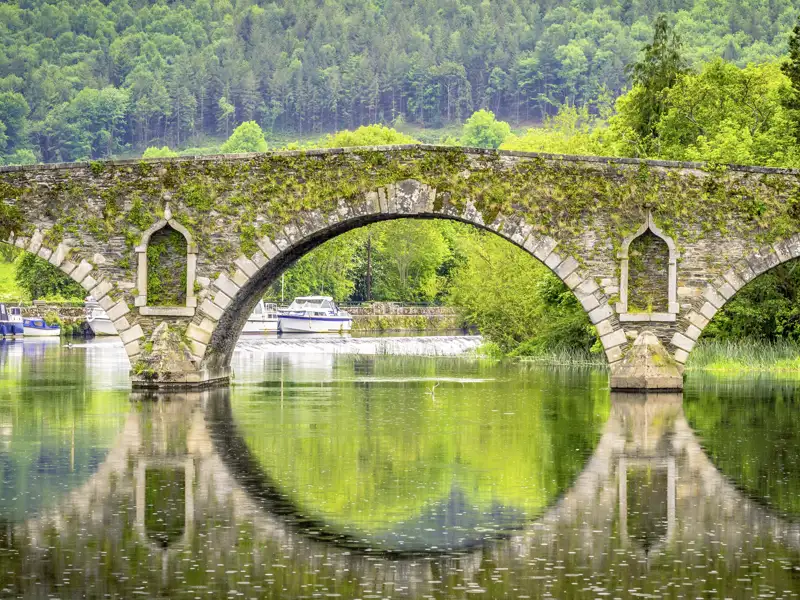 Auf unserer Rundreise durch Irland machen wir halt in Kilkenny, das für seine zahlreichen lebendigen Pubs ebenso berühmt ist wie für seine Gebäude. Hier eine historische Steinbrücke mit moosbewachsenen Bögen und Booten im Fluss.