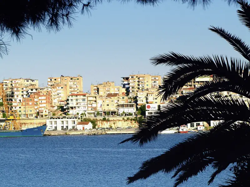Palmen rahmen den Blick auf die Gebäude der Küstenstadt Saranda und den Hafen ein. Von hier starten wir unsere Tagesausflüge nach Gjirokastra und Butrint.